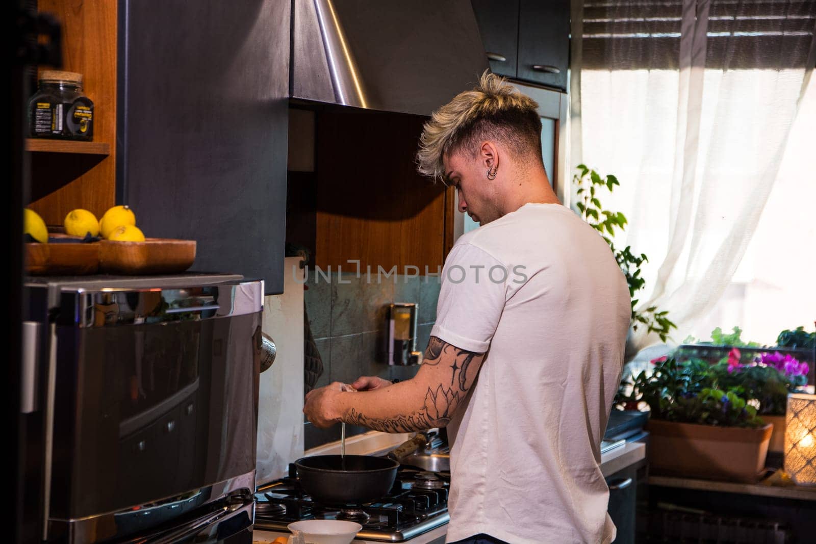 A handsome young man with blue eyes is cooking in a stylish kitchen. He is stirring ingredients in a bowl while surrounded by fresh vegetables and kitchenware. Sunlight streams through the window.