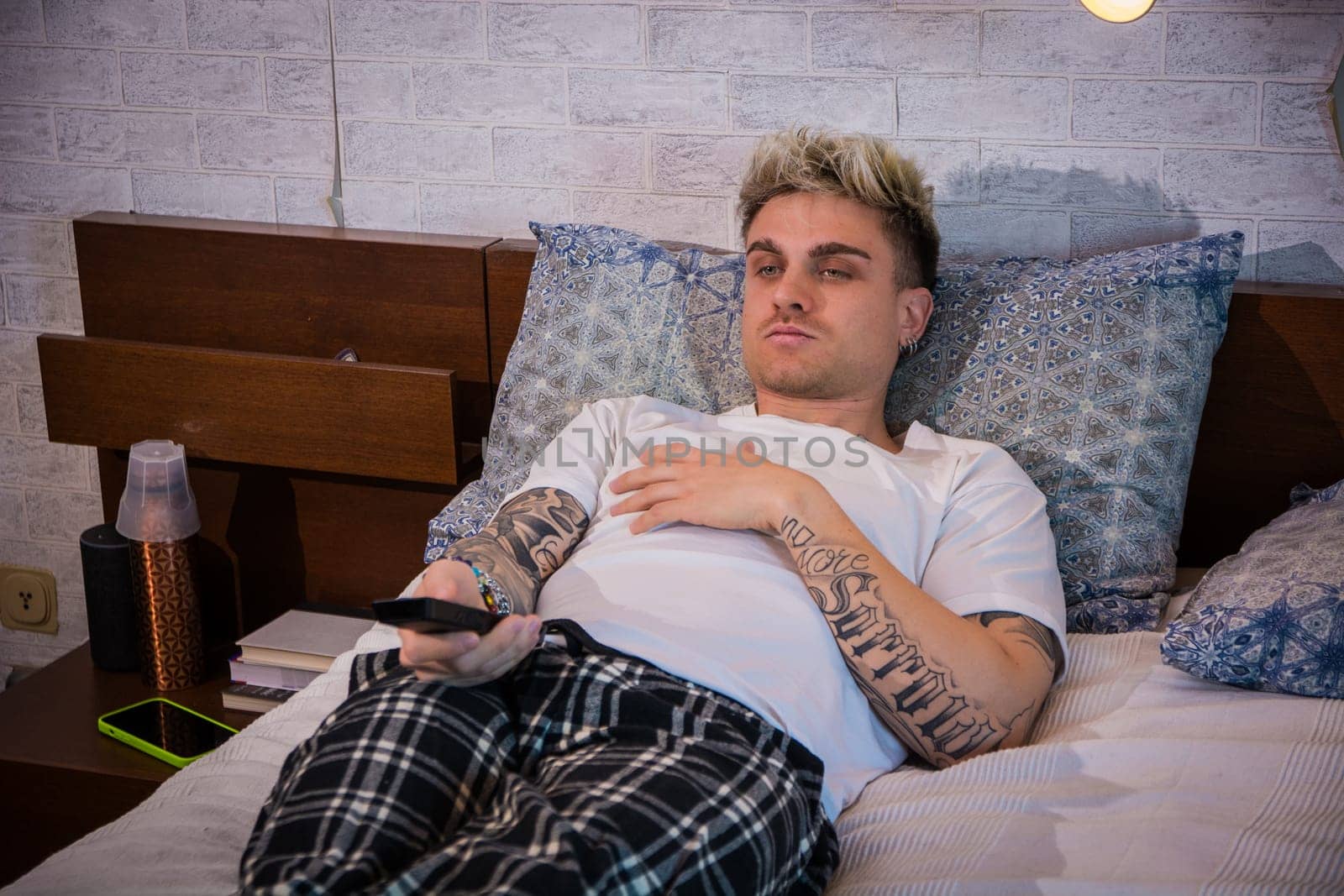 A young man with blond hair and blue eyes rests on a bed, dressed casually in a t-shirt and plaid pants. He focuses on the television, enjoying a calm evening at home.