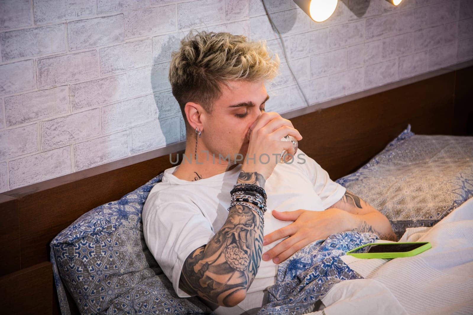 A young man with blond hair and blue eyes is seated on his bed, sipping from a glass. He has visible tattoos on his arms and appears relaxed in a cozy bedroom setting.