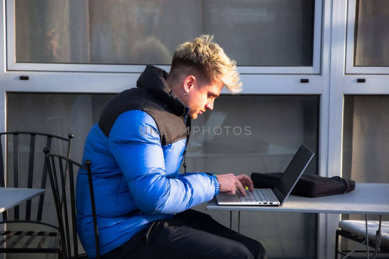 A young man with blond hair sits at a table concentrated on his laptop. Dressed in a blue jacket, he works outdoors in a modern environment, enjoying the bright daylight.