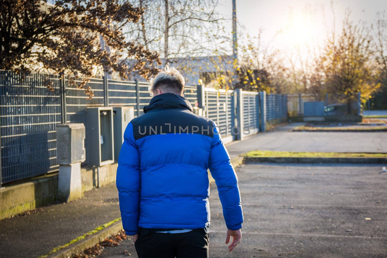 A young man with blond hair and blue eyes walks away in a blue and black jacket. The sun sets behind him, creating a warm atmosphere in a quiet urban area.