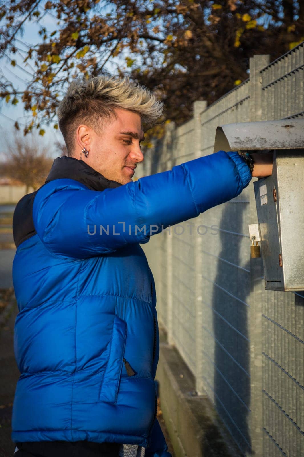 A young man with blond hair and blue eyes is checking his mailbox while wearing a blue jacket. It is a sunny day, and the setting appears to be an urban environment.