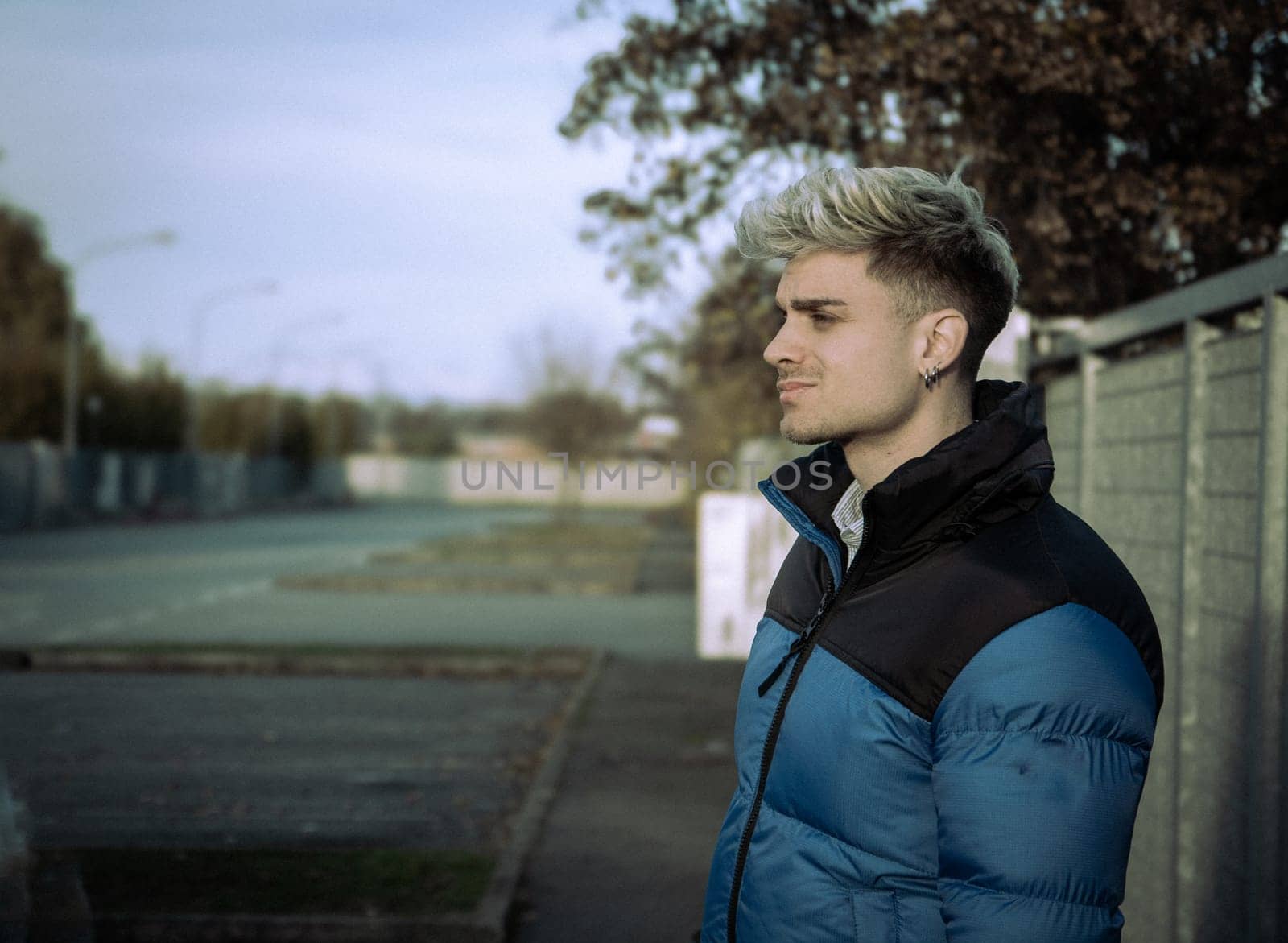 A young man with striking blond hair and blue eyes stands outdoors, wearing a blue jacket. He exudes confidence as he gazes thoughtfully into the distance on a tranquil afternoon.