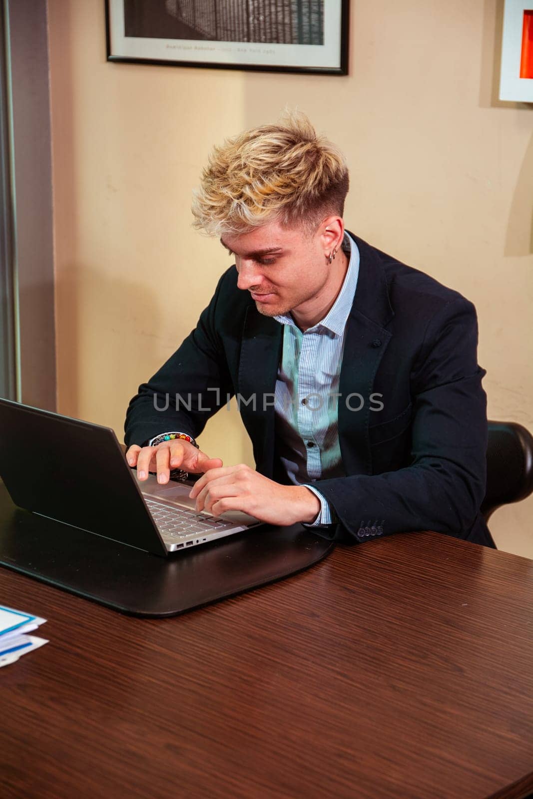 A handsome young man sits at a desk focused on his laptop. He has blond hair and blue eyes, dressed in a suit, clearly engaged in his work environment during daylight hours.
