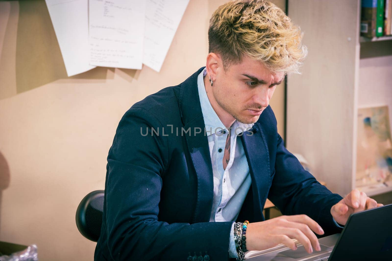 A handsome young man with blond hair and blue eyes is deeply engaged in his work at a desk. He is focused on his laptop, displaying determination in a contemporary office environment.