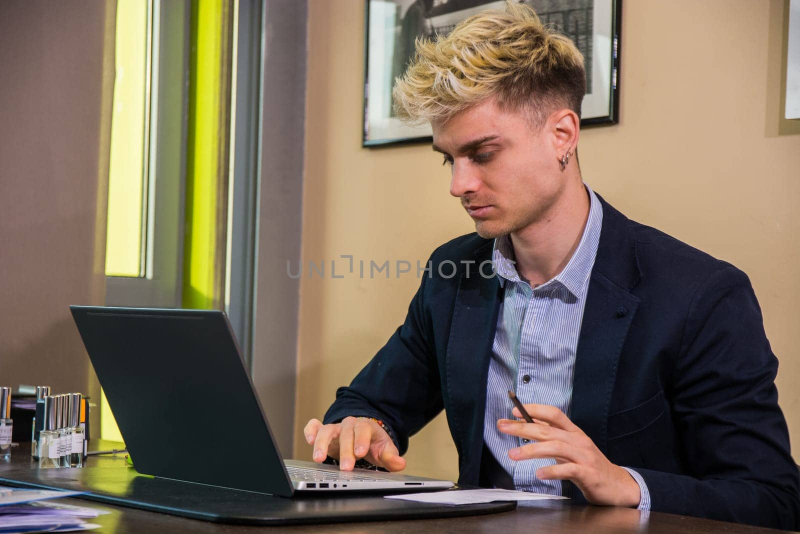 A handsome young man with blond hair and blue eyes is focused on his laptop, dressed in a smart blazer while seated at a contemporary office desk filled with stationery.