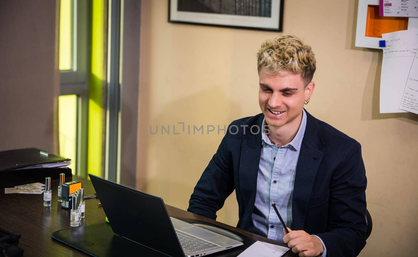 A young man with attractive features sits in a sleek office, focused on his laptop screen. He wears a navy blazer and smiles as he holds a pen, surrounded by papers and office supplies.