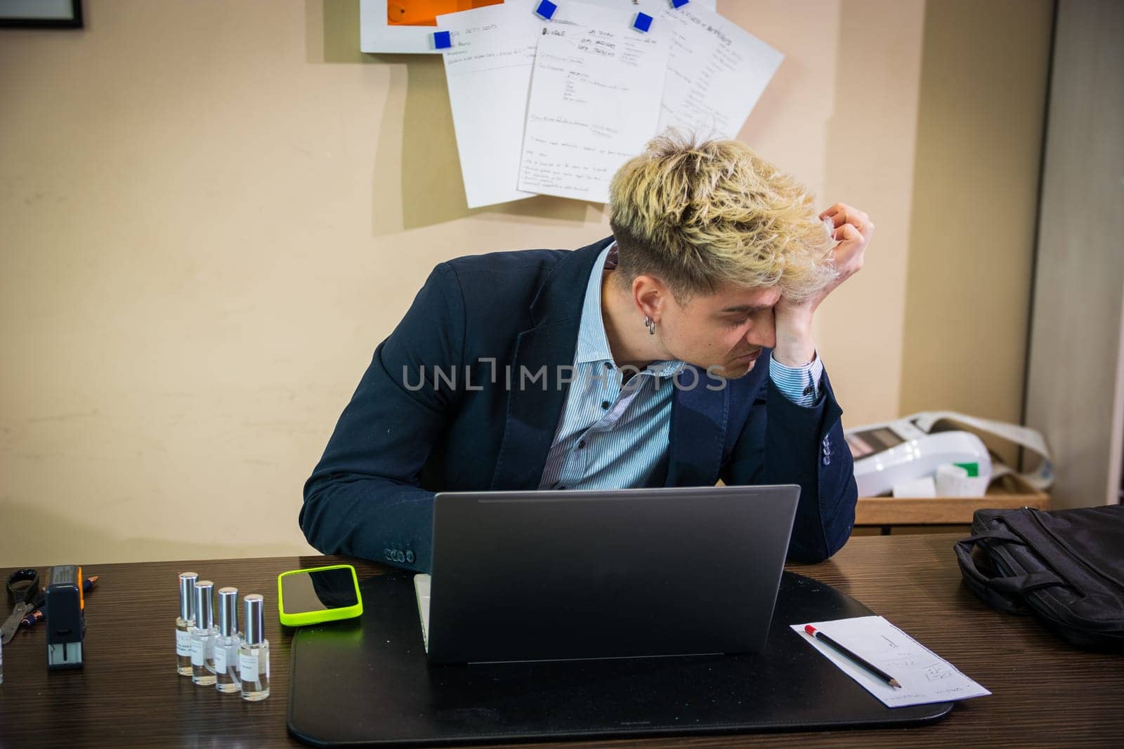 A young man with blond hair and blue eyes sits at a desk, looking concentrated as he works on his laptop. Papers and equipment surround him in the bright, contemporary office.