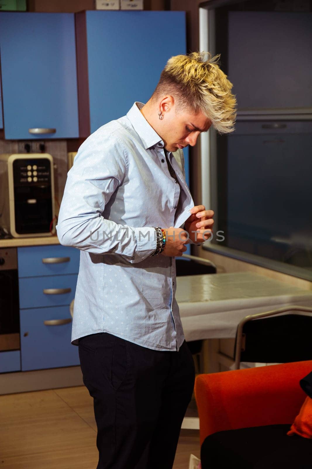 A young, attractive man with blond hair and blue eyes is adjusting his button-up shirt in a contemporary kitchen. The space features blue cabinets and a stylish design that complements his look.