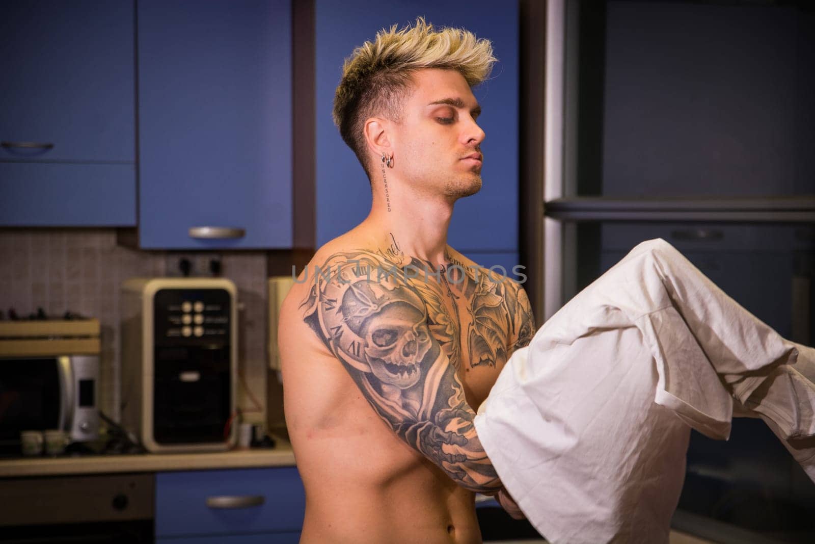 A young man with blond hair and striking blue eyes stands in a sleek kitchen. He holds a white cloth, showing off his tattoos, as he concentrates on his task in the warm evening light.