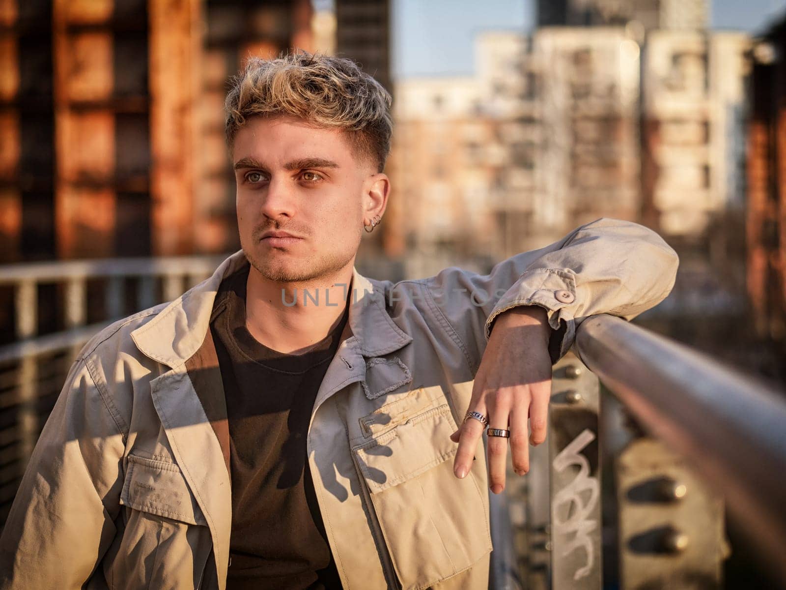 A young blond man with blue eyes leans against a railing in an urban setting at dusk. His stylish outfit and relaxed posture convey confidence as the city lights begin to glow.