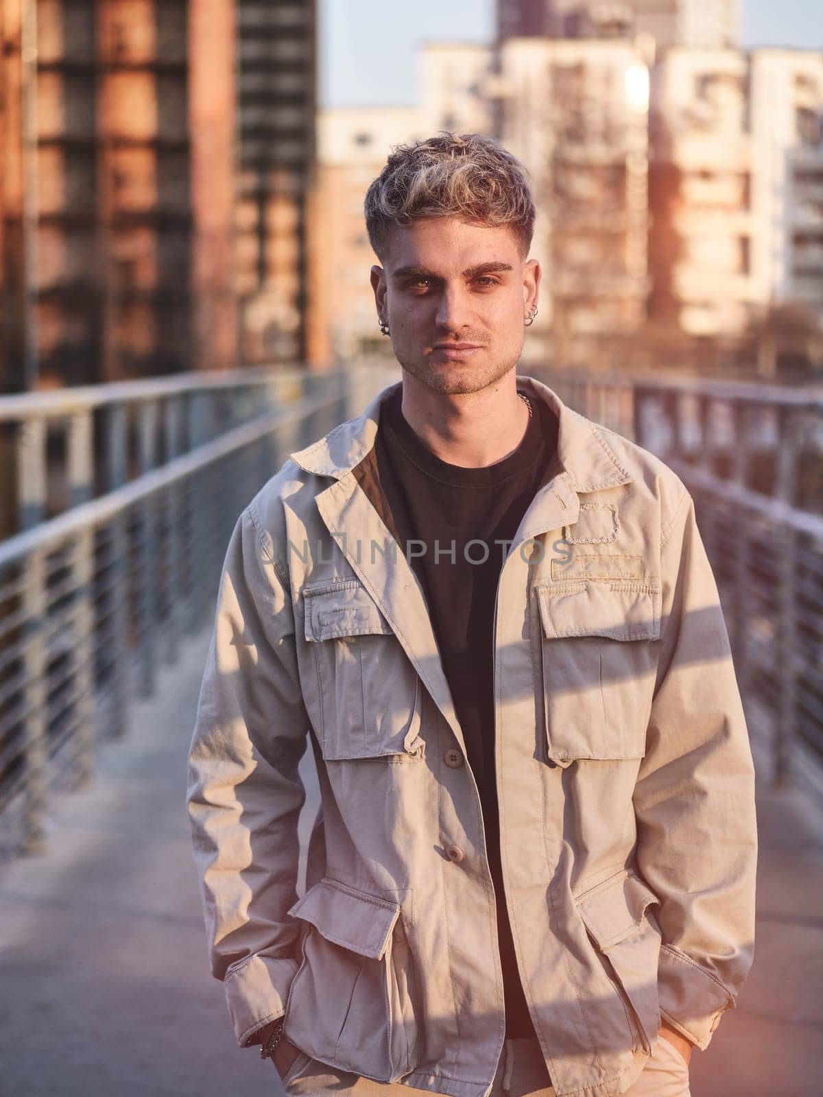 A handsome young man with blond hair and blue eyes stands on a contemporary urban bridge. The warm glow of sunset highlights his features, enhancing the stylish look of his casual outfit.