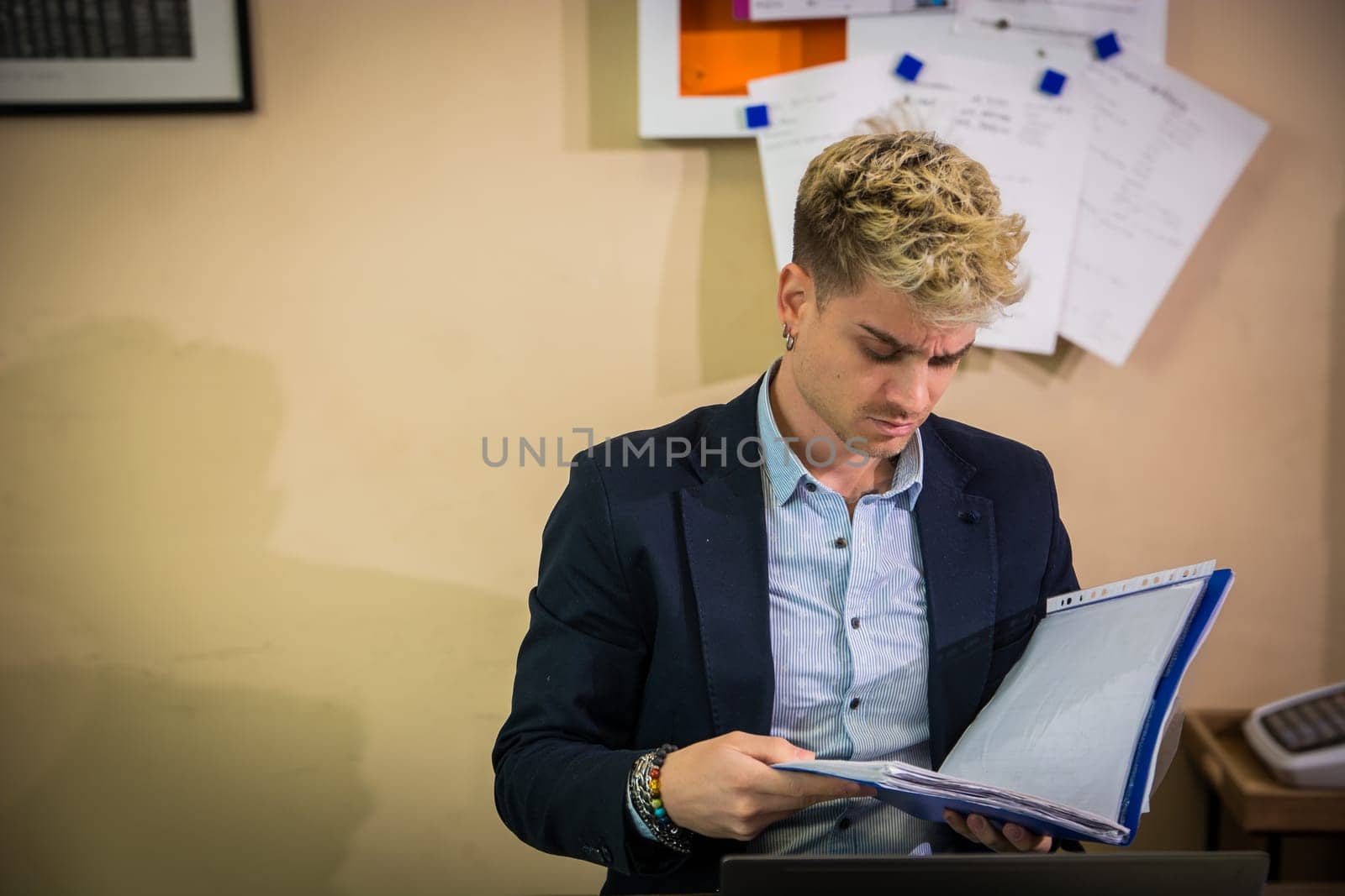 A young man with blond hair and blue eyes examines a folder filled with documents. He appears focused and engaged, seated at a desk in a contemporary office setting.