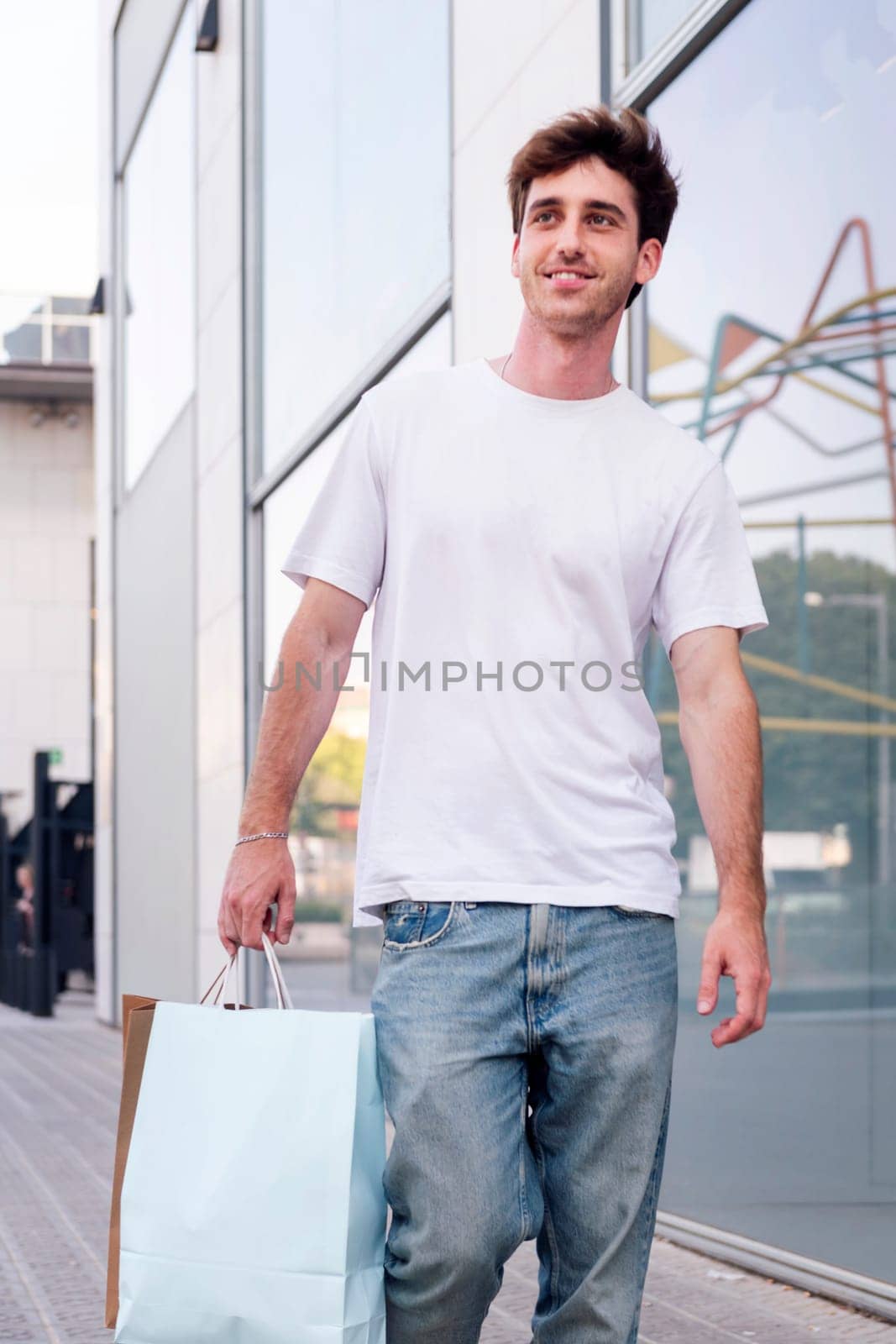 young adult man in casual clothes walking down a sidewalk near of a mall carrying shopping bags, concept of youth and urban lifestyle, copyspace for text
