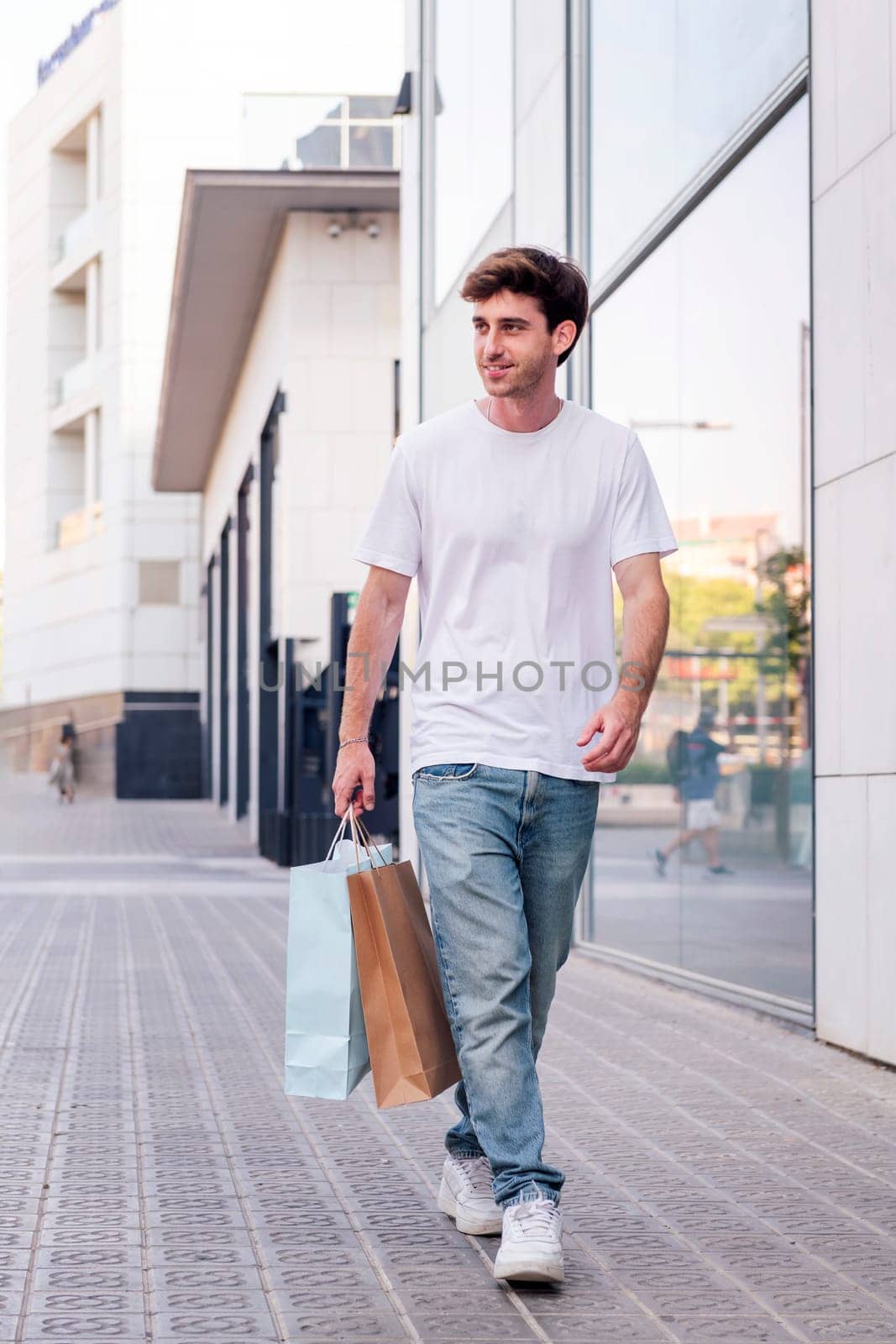 young adult man in casual clothes walking down a sidewalk near of a mall carrying shopping bags, concept of youth and urban lifestyle, copyspace for text
