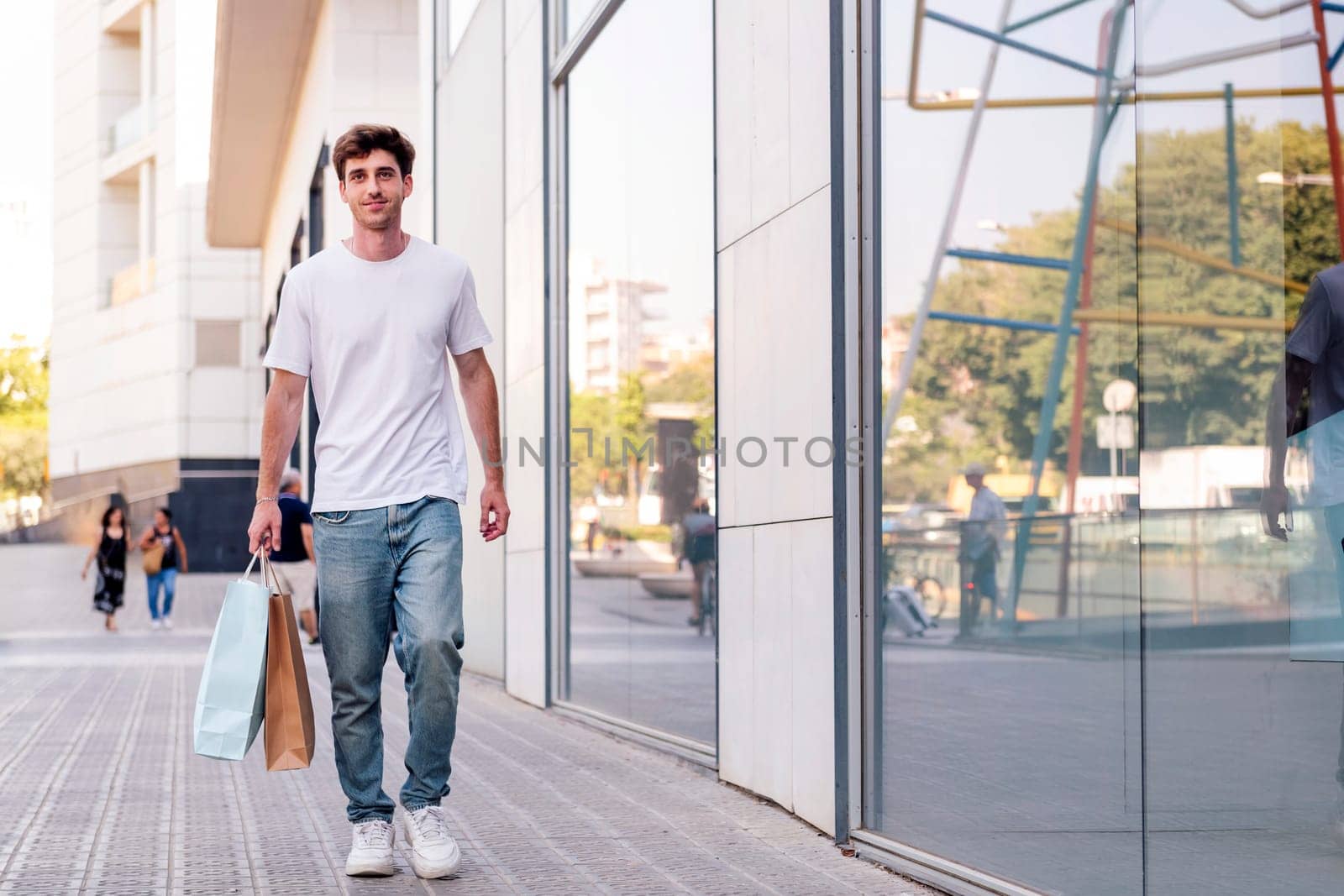 young adult man in casual clothes walking down a sidewalk near of a mall carrying shopping bags, concept of youth and urban lifestyle, copyspace for text