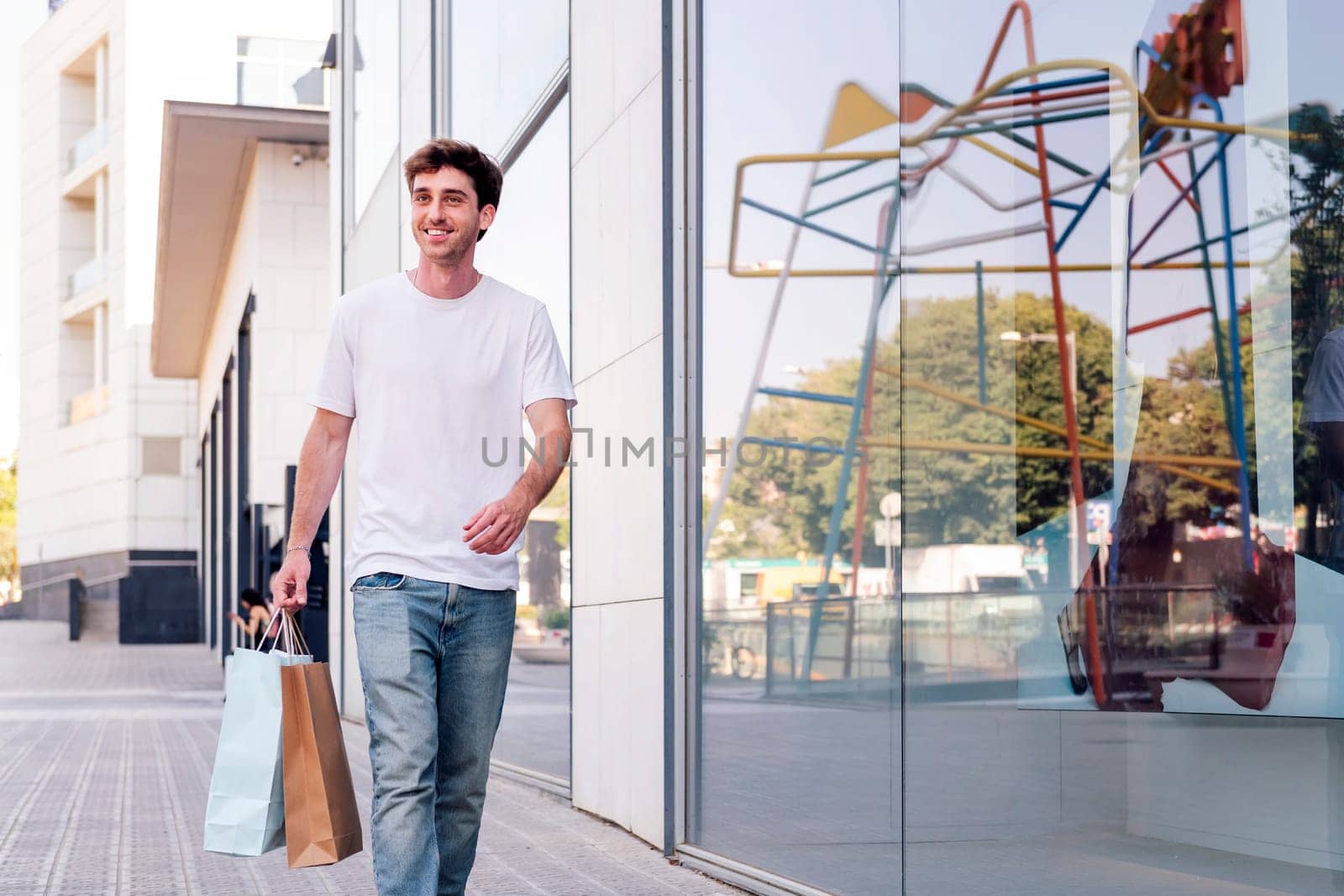 young adult man in casual clothes walking down a sidewalk near of a mall carrying shopping bags, concept of youth and urban lifestyle, copyspace for text