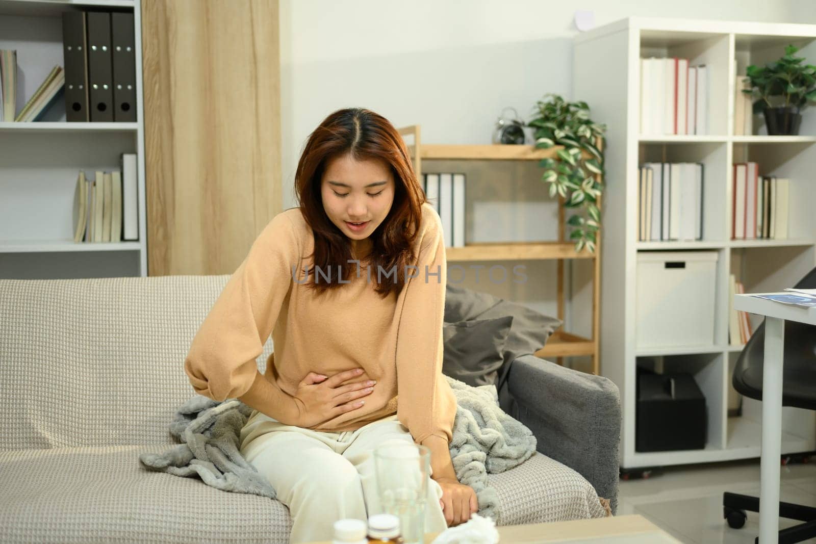Asian woman suffering from stomach pain while sitting on sofa. Period cramps or gastrointestinal issues.
