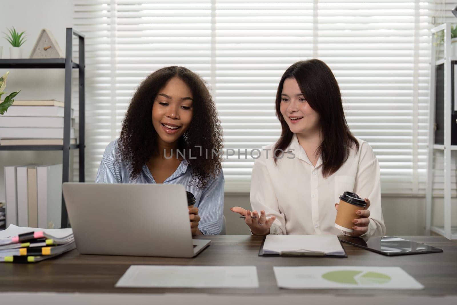 Two friends at work laugh and discuss ideas over coffee and a laptop, showcasing a positive and encouraging collaborative atmosphere.