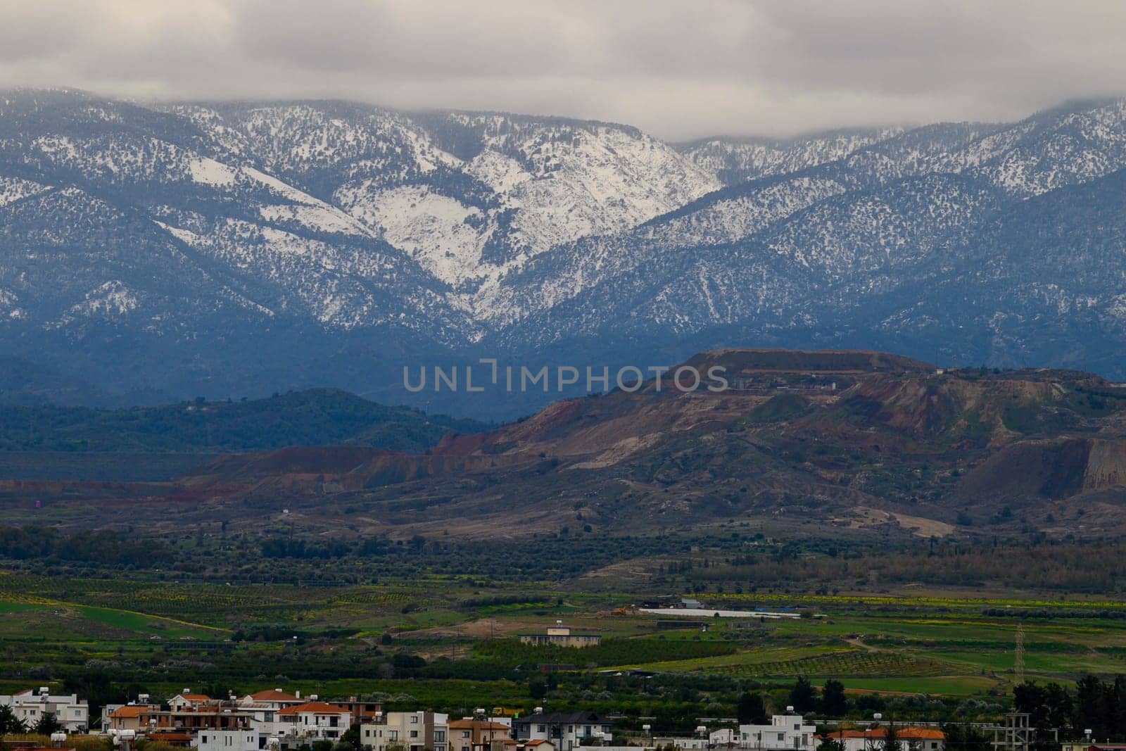 Majestic snow-capped peaks rise high above lush green vineyards and small farms, creating a stunning backdrop on a chilly evening filled with clouds and tranquility.