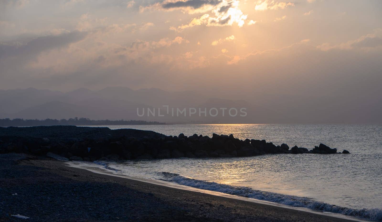 Soft sunlight filters through the clouds, casting a warm glow on the sea as gentle waves lap against a stony shore, creating a serene atmosphere at dusk by the coast.