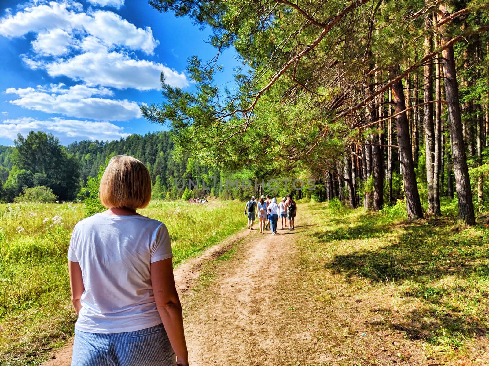 A woman walks ahead of a group on a forest path under blue skies, surrounded by tall trees and lush greenery, enjoying nature.