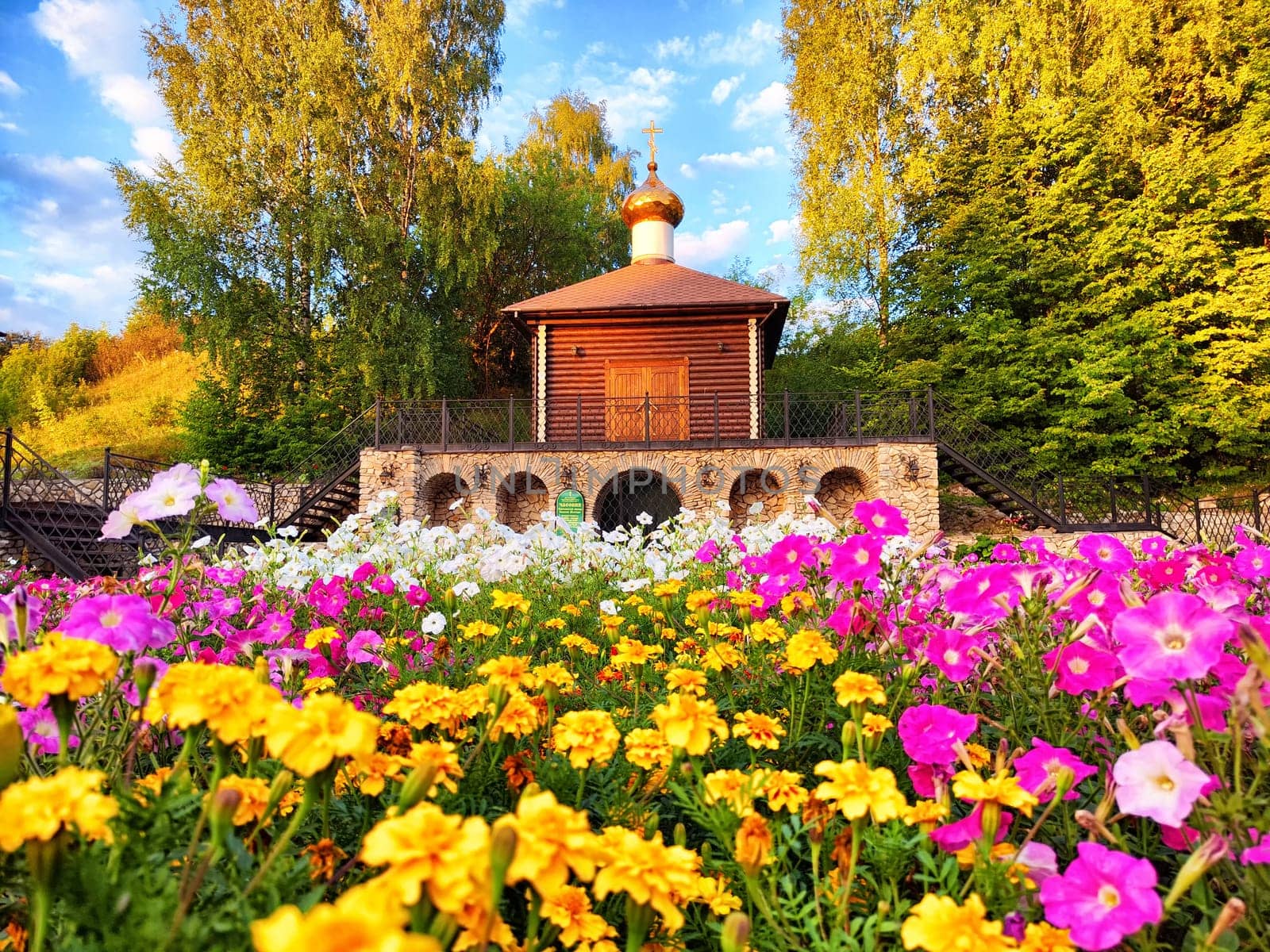 Vibrant flowers create a stunning foreground while a quaint wooden building with a golden dome stands among the trees under a bright sky.