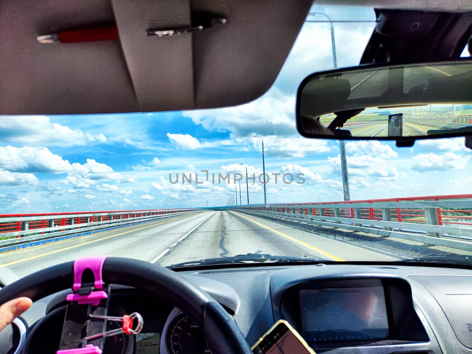 A hand grips the steering wheel while driving across a long bridge under a clear blue sky, with vibrant clouds above and bright sunlight.