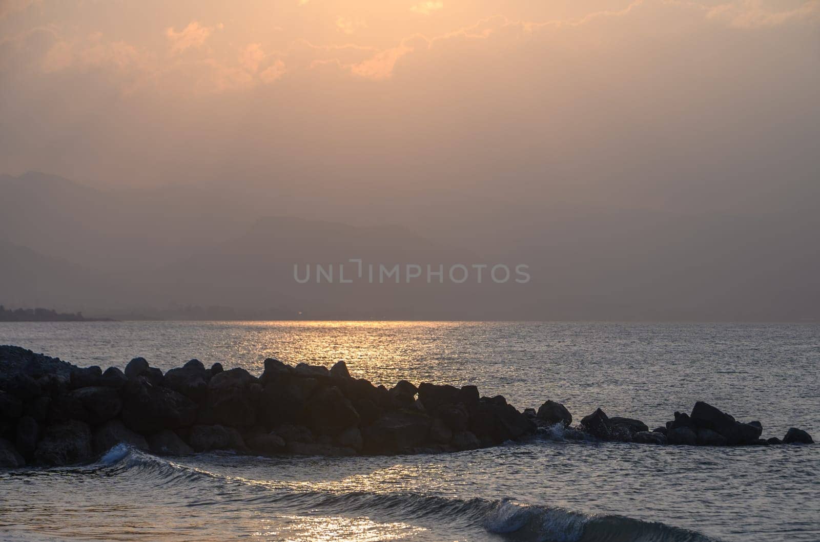Golden hues blanket the horizon as the sun sets over tranquil waters. Waves gently lap against the rocky shore, creating a peaceful environment at dusk.