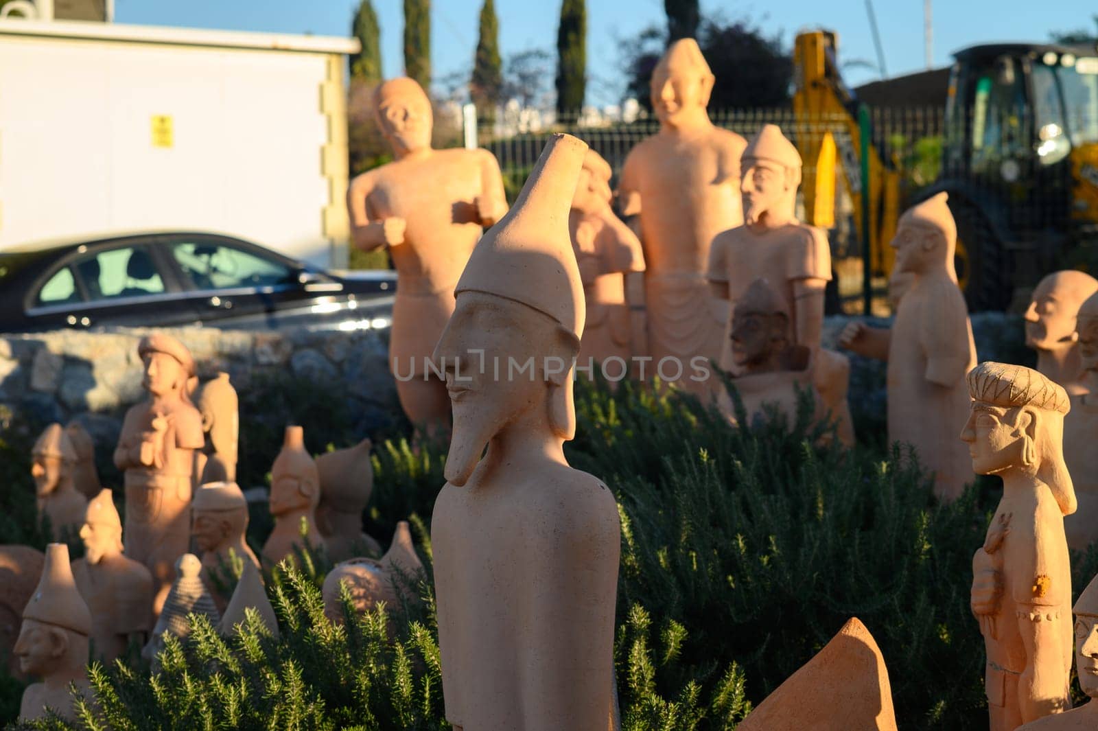 A diverse array of clay sculptures stands proudly in a sunlit outdoor space, showcasing various artistic styles and figures among greenery. The warm light highlights their intricate details.