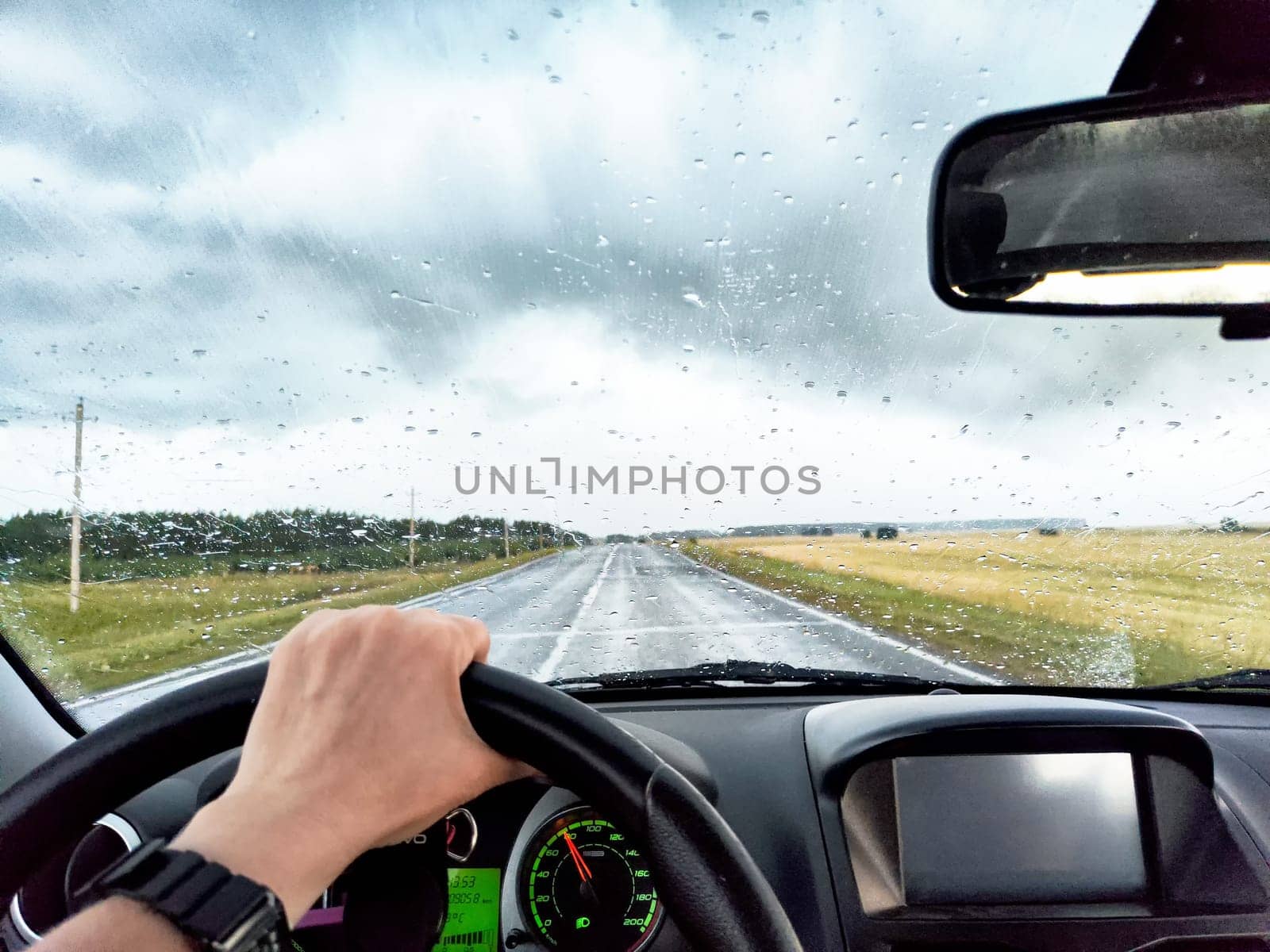 A person drives a car down a wet road with raindrops on the windshield, flanked by open fields and overcast skies.