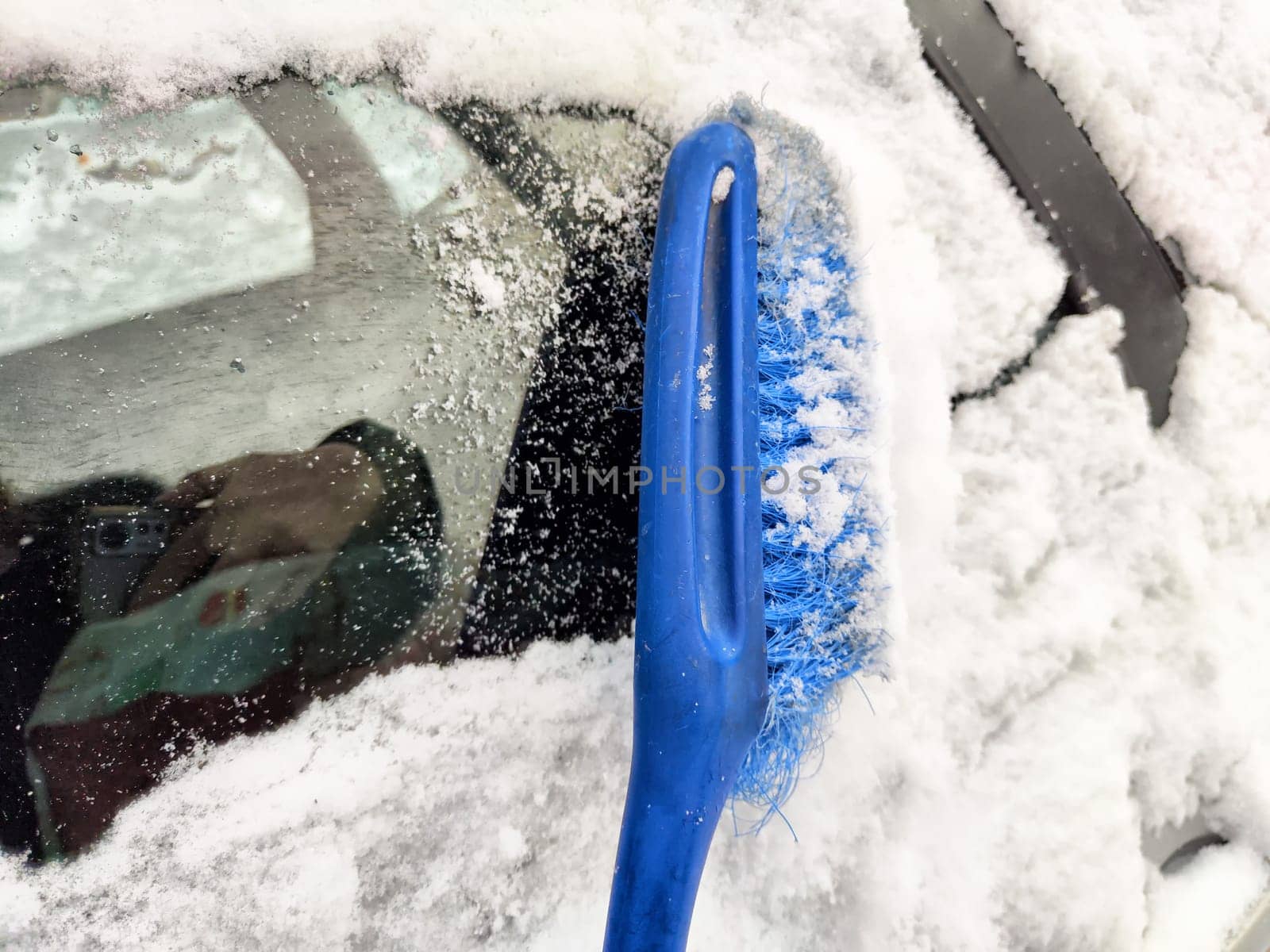 Using a brush to remove snow from a frozen car window on a cold winter morning in a snowy landscape.