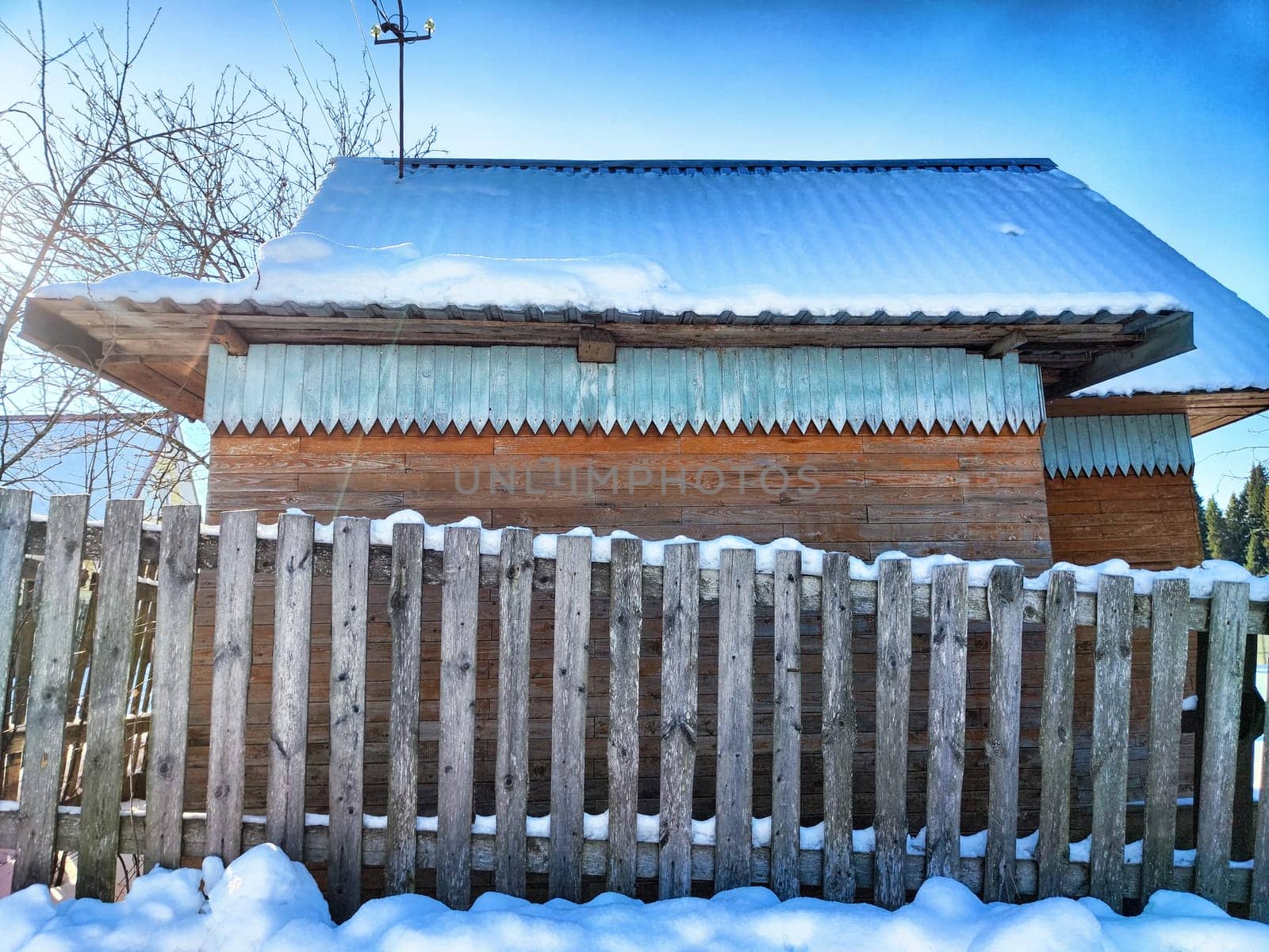 A rustic wooden house stands quietly amidst snow, surrounded by a weathered wooden fence under a clear blue sky during winter.