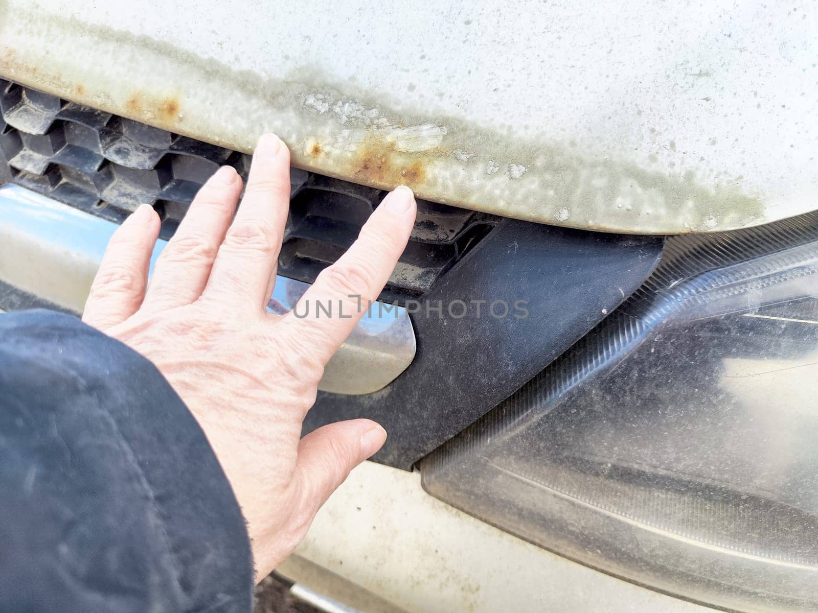 A person inspects the corroded front grille of an older vehicle while standing in a rural environment.