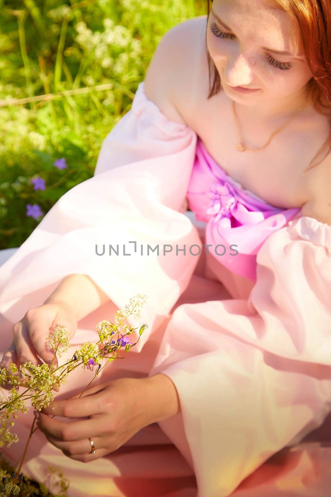 A young woman gently collects wildflowers while seated in a brightly lit meadow, adorned in a flowing pink dress during a warm afternoon.