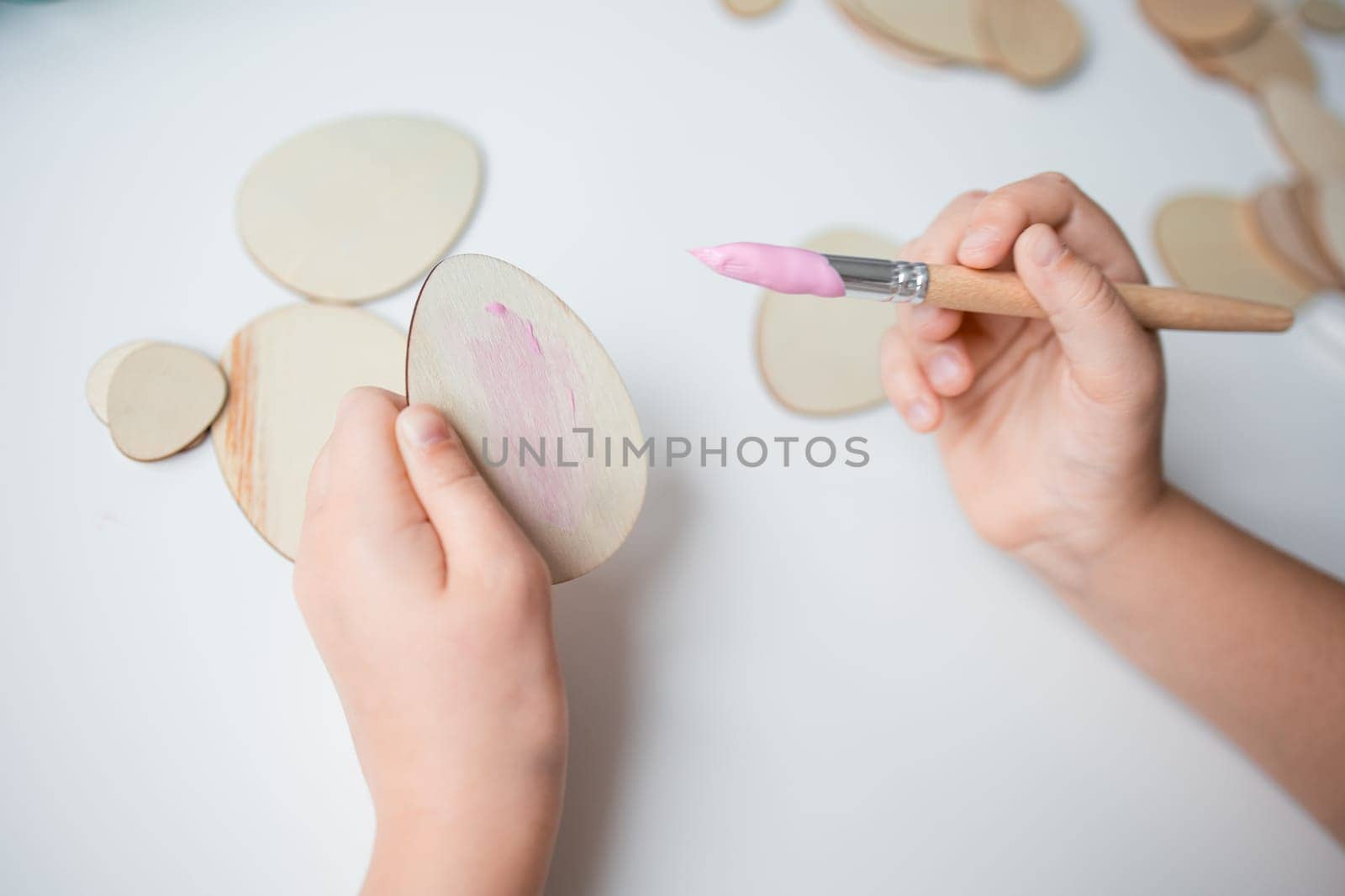 Child painting Easter wooden egg shapes with pink paintbrush on white surface.