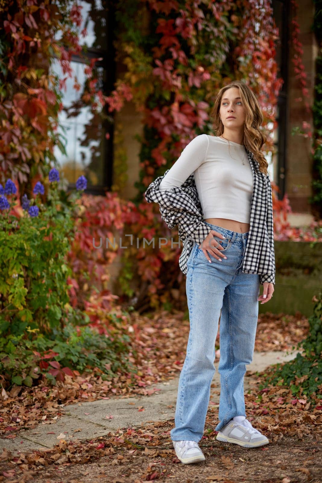 Stylish young woman in trendy autumn outfit posing confidently in a park with vibrant fall foliage, wearing a black-and-white plaid shirt over a white crop top, paired with blue jeans and white sneakers, exuding casual yet fashionable vibes.