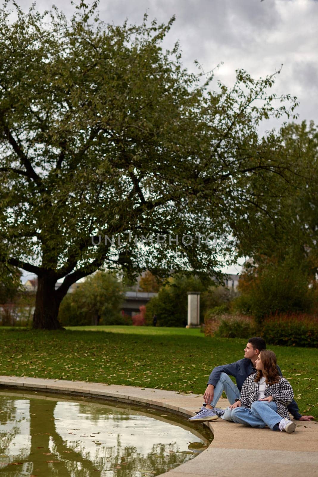 Autumn Serenity: Couple Relaxing by Reflective Pond Amidst Vibrant Fall Foliage and Ivy-Covered Architecture. Tranquil Garden Scene Captures Intimate Moment Against Backdrop of Colorful Autumn Leaves and Mirrored Water Surface. This enchanting autumn scene portrays a couple sharing a peaceful moment by a serene pond, surrounded by the rich hues of fall foliage. The pair sits comfortably on the grass, leaning against each other in quiet companionship as they gaze upon the reflective water. Vibrant red and orange leaves adorn the surrounding trees, creating a warm and inviting atmosphere. An ivy-covered building in the background adds a touch of timeless charm to the landscape. The pond's mirror-like surface perfectly captures the reflection of the couple and the colorful foliage, doubling the visual impact of autumn's beauty. This image embodies the essence of autumn tranquility, making it ideal for themes of romance, relaxation, and the changing seasons.
