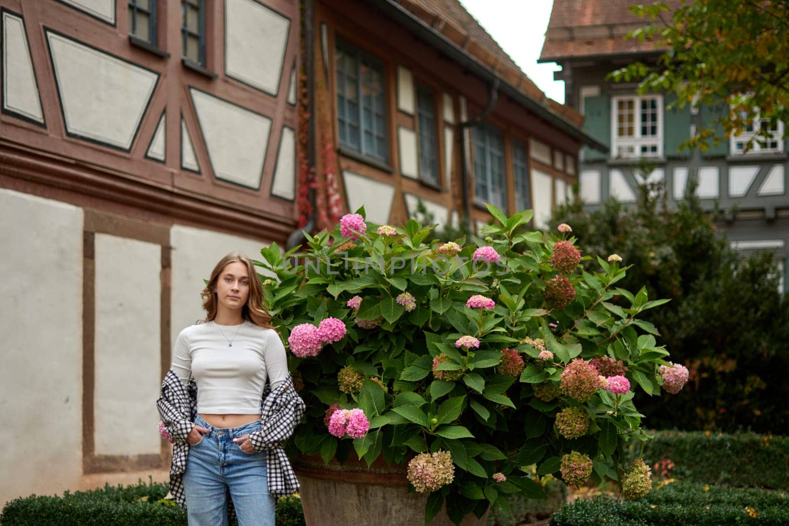 This image features a young woman in her early twenties posing joyfully in a picturesque European garden. She is wearing a casual outfit, including a white crop top, an unbuttoned checkered shirt, and loose blue jeans, embodying a relaxed and stylish aesthetic. Her pose is playful and confident, with one arm stretched upward, conveying a sense of freedom and happiness. She is positioned next to a large planter filled with blooming hydrangeas in vibrant shades of pink, adding a vivid touch of color. The background showcases traditional European half-timbered houses with brown frames and white walls, complemented by green window shutters and creeping ivy, emphasizing the charm of a rural village setting. The scene is bathed in soft natural light, highlighting the lush greenery and creating a warm, inviting autumn atmosphere. Perfect for themes such as travel, lifestyle, fashion, nature, and European culture, this photograph captures the beauty of everyday moments in a serene countryside setting. Young woman posing with hydrangeas in a European garden, surrounded by half-timbered houses and autumn foliage, expressing joy and elegance.