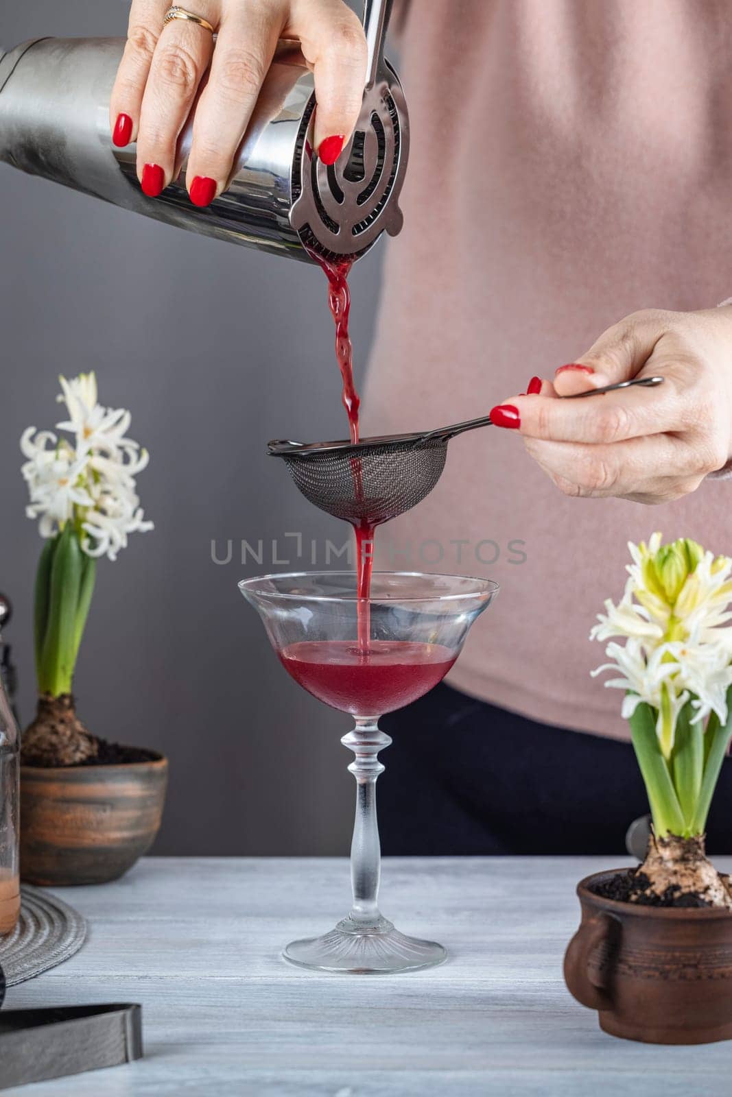 Bartender filtering red cocktail from shaker through strainer into a coupe glass, white hyacinth flowers on a gray background