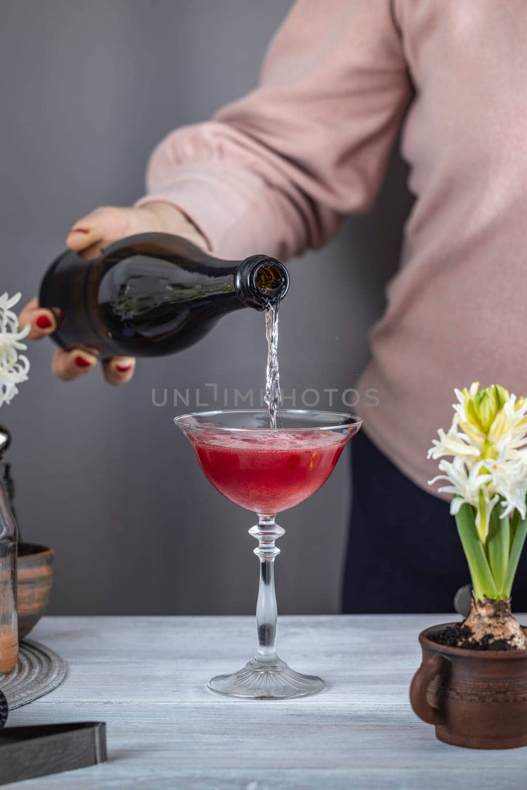 Bartender pouring sparkling wine into a cocktail glass with red liquor, ice, and flower decorations for a refreshing drink