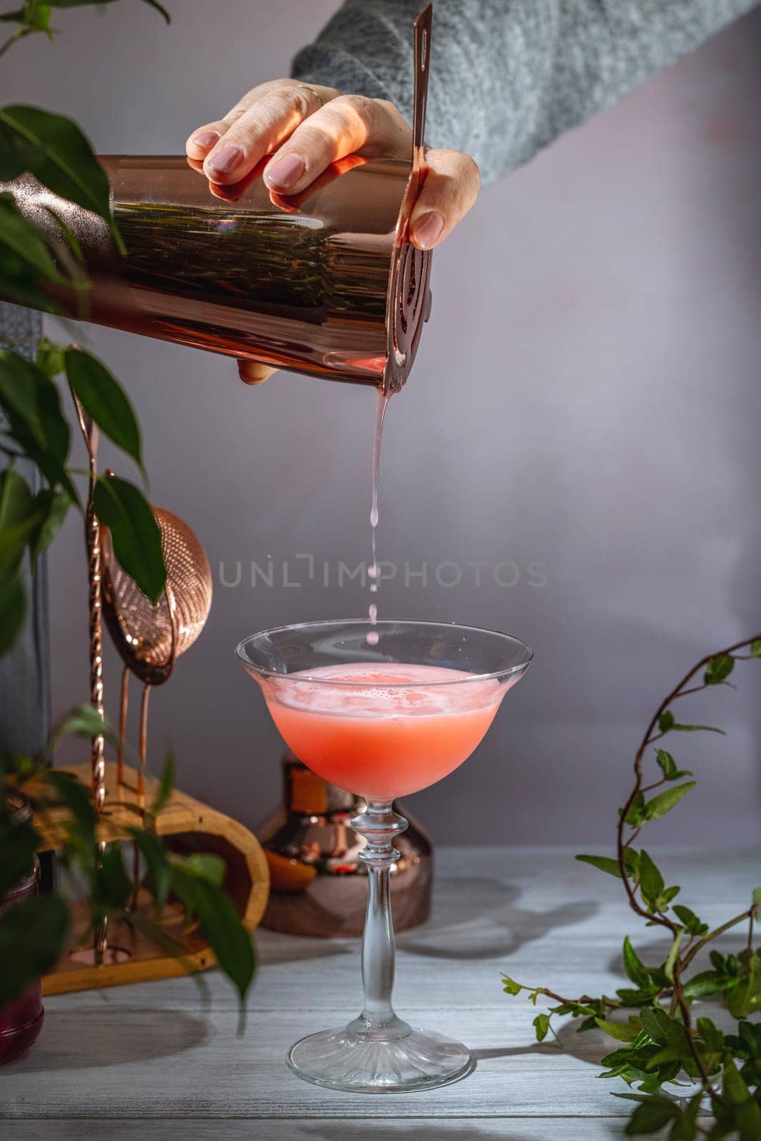 Bartender pours a vibrant pink cocktail from a copper shaker into a stemmed glass, surrounded by bar tools and lush greenery