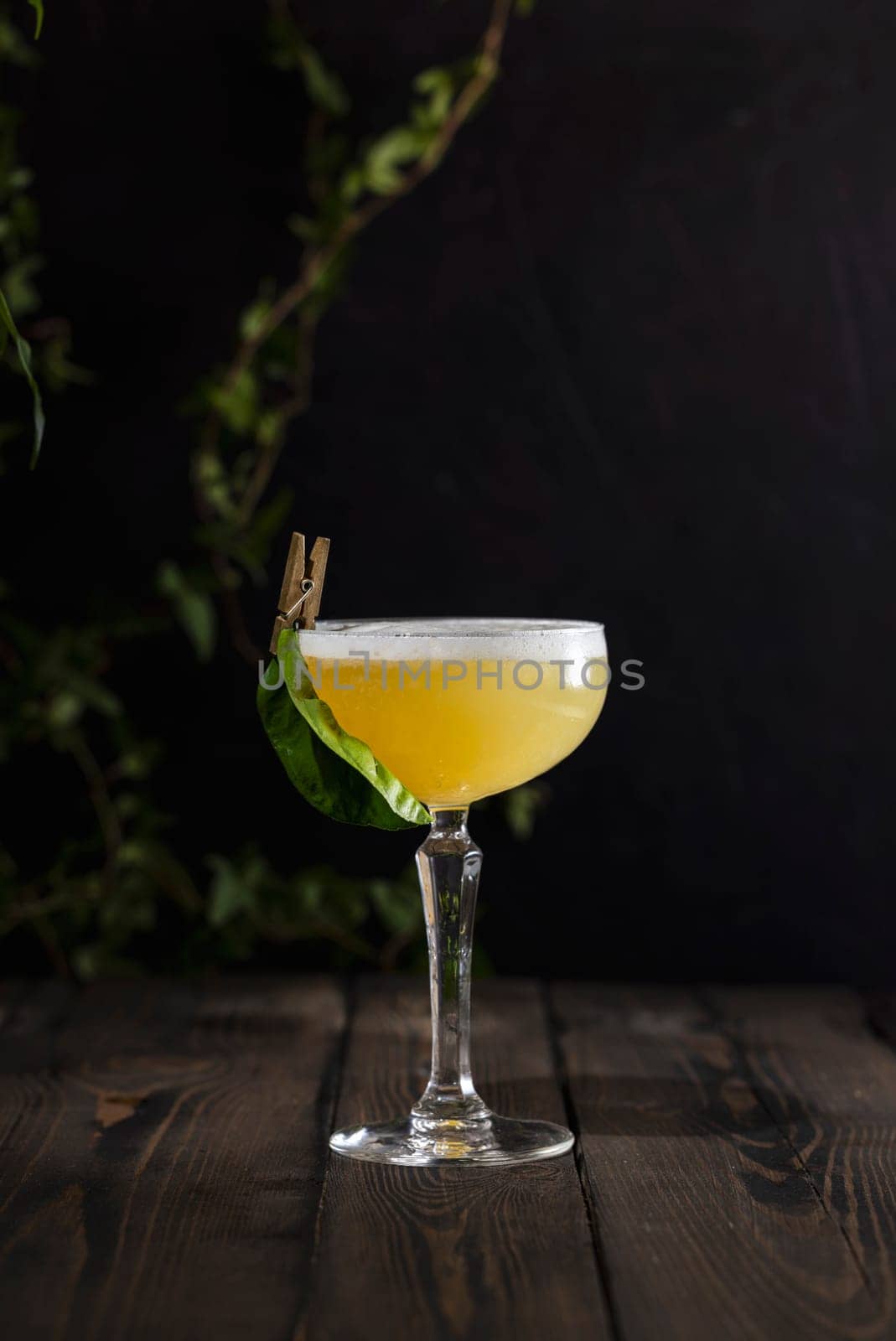 Bright yellow cocktail in a stemmed glass, garnished with a fresh green leaf and clothespin, resting on a dark wooden table