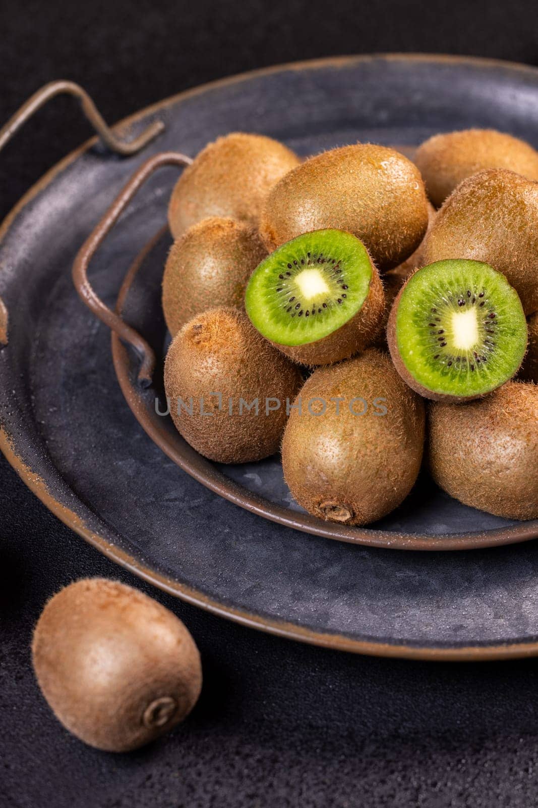 Kiwi fruits are shown whole and cut open, resting on a dark, rustic metal tray, highlighting their vibrant green flesh and fuzzy brown skin