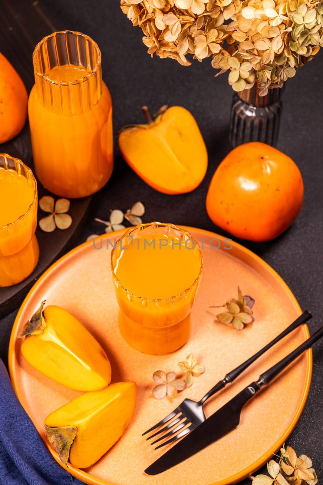 Glass of refreshing persimmon juice served with ripe persimmon fruits on a plate, decorated with dried flowers, creating a vibrant autumnal table setting