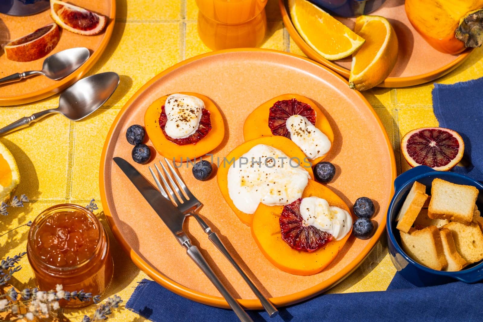 Delicious persimmon dessert with ricotta cheese, blood orange slices and blueberries, served on a vibrant table with orange juice, jam, and melba toast