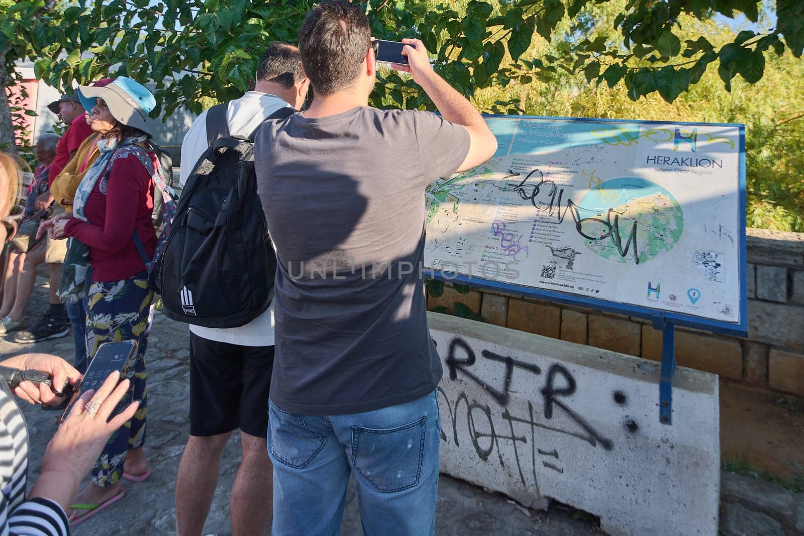 Crete.Greece - april 02, 2025: A group of tourists looking at a map of Heraklion outdoors, engaging with city details and planning their visit, with visible trees and a stone wall in the background.