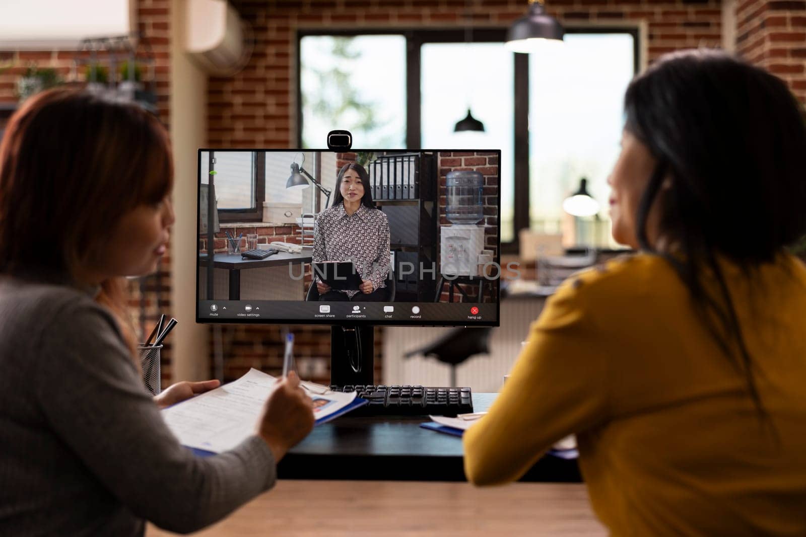 HR manager on computer screen, conducting a virtual meeting with her employees, discussing different job applicants. Female office workers seated at desk with paperwork, on a video call with employer.