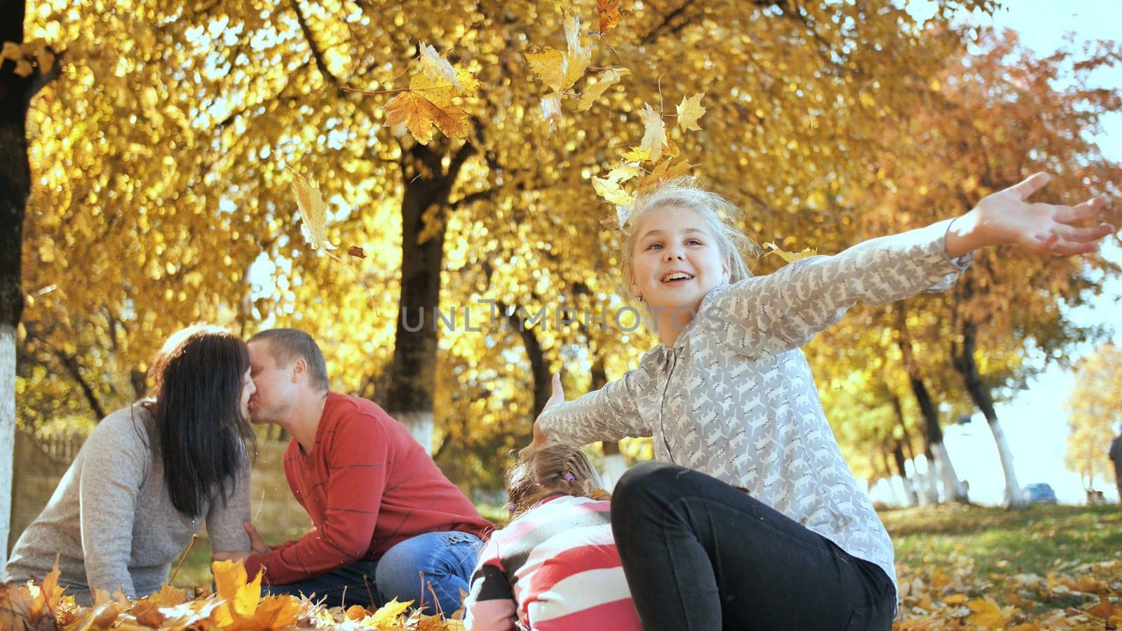Happy family outdoors playing with fallen leaves in sunny autumn weather. Parents kiss in the background