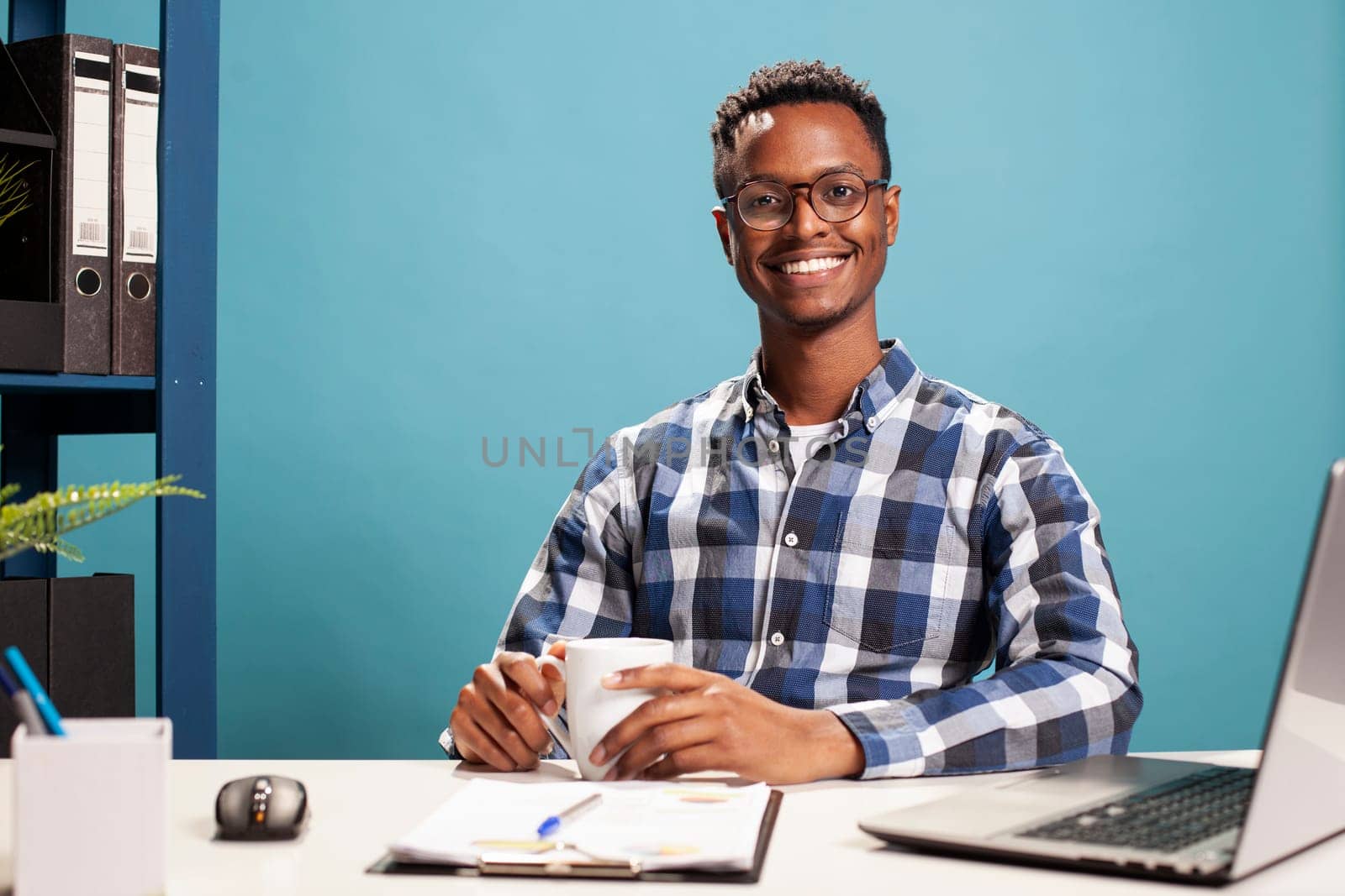 Young black man with coffee cup in his hands, is sitting at his desk and smiling at the camera. African american male freelancer seated at table with clipboard and a laptop, ready for work.